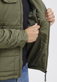 Groene gewatteerde jack met een rits en drukknoop, met een binnenzak en een gladde voering. De mouw heeft ribbelmanchetten en een rechte zoom.