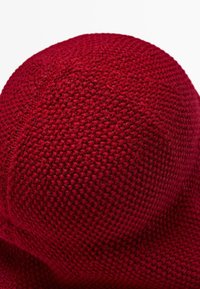 Close-up of a deep red knit beanie showing detailed textured stitch patterns and seam lines on a curved surface.