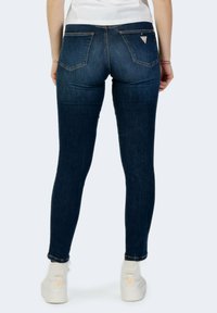 Jeans skinny azul oscuro con una textura suave, que cuentan con dos bolsillos traseros y una pequeña etiqueta de logo cerca de la cinturilla. Combinados con zapatillas blancas.