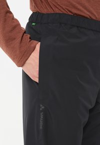 Pantaloni impermeabili neri con cintura elastica, dotati di una tasca laterale e del logo "Vaude" visibilmente esposto sulla parte bassa della gamba.