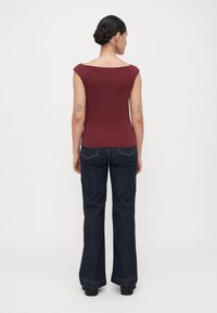 Mujer con moño trenzado oscuro que lleva una blusa sin mangas borgoña y jeans anchos de pierna oscura, de pie de frente a un fondo claro y sencillo.