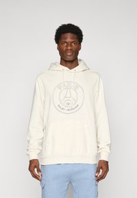 Muž oblečený v off-white mikine Paris Saint-Germain a svetlomodrých cargo nohaviciach, stojaci na pozadí v jednoduchom svetlošedom tóne.