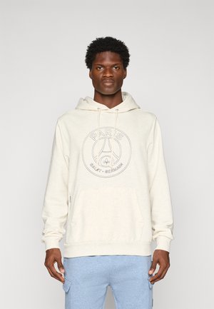 Uomo che indossa un hoodie off-white del Paris Saint-Germain e pantaloni cargo azzurri chiari, in piedi contro uno sfondo grigio chiaro uniforme.