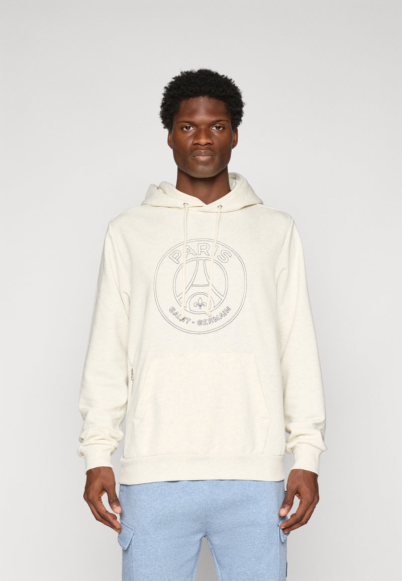 Muž oblečený v off-white mikine Paris Saint-Germain a svetlomodrých cargo nohaviciach, stojaci na pozadí v jednoduchom svetlošedom tóne.