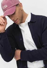 Marinblå ribbad stickad cardigan med knappar, bärs över en vit skjorta med krage. En vinröd keps med "SUN DAY" broderat justeras.