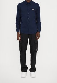 Camicia azzurra navy con bottone e maniche lunghe, con una toppa sul petto, abbinata a pantaloni cargo neri con tasche laterali e sneakers bianche.
