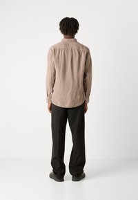 Camicia di lino marrone vista da dietro, con un orlo curvo, polsini abbottonati e un fit rilassato, abbinata a pantaloni neri e scarpe.