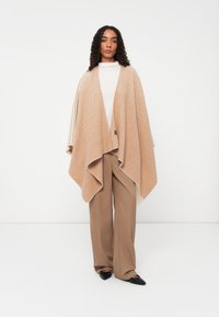 Beige ullponcho med et teksturert design, som har en drapert silhuett, asymmetrisk kant og et subtilt stripemønster.
