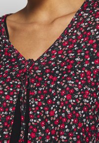 Imagen en primer plano de un pecho que lleva una blusa negra con un cárdigan floral que presenta pequeñas flores rojas, blancas y rosas atadas en el cuello.