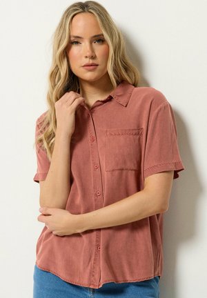 Blondynka z długimi włosami, ubrana w różową, krótko rękawową koszulę zapinaną na guziki z kieszenią na piersi, pozująca na białym tle.