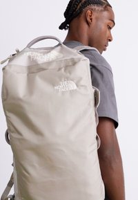 Mladý muž s copánky, oblečený v šedém tričku, nese na zádech velký béžový batoh The North Face.
