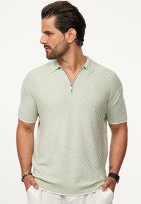 Kurzärmliges Polohemd in Hellgrün mit einem strukturierten Muster, Reißverschlusskragen und gerippten Bündchen. Der Stoff scheint weich und leicht zu sein.