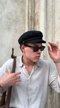 Czarna bawełniana czapka z plecionym wykończeniem, czarne okulary przeciwsłoneczne, lekka lniana koszula i brązowa skórzana torba na ramię na tle teksturowanym w jasnym kolorze.