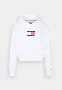 Vit cropped hoodie gjord av mjukt material, med en marinblå lapp med "TOMMY JEANS" och en amerikansk flaggdesign på framsidan. Justerbara snoddar.
