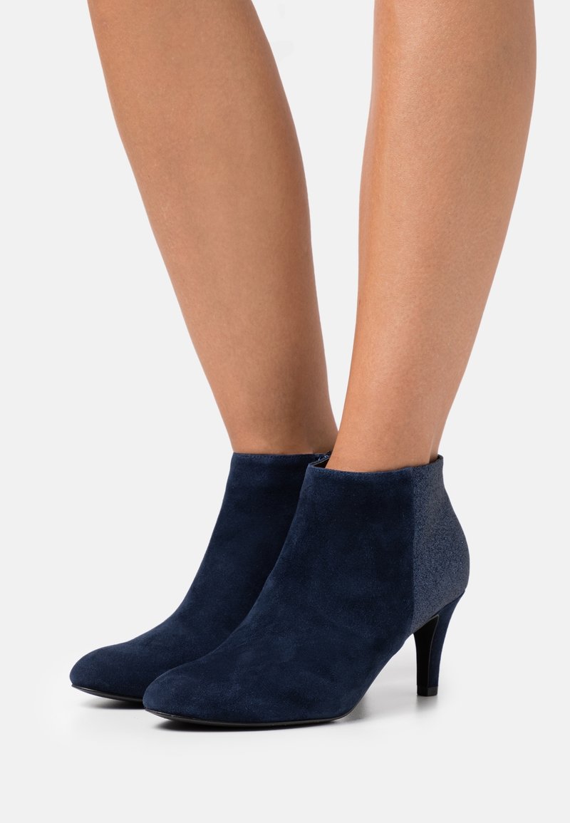Femme portant des bottines en daim bleu marine à petits talons, debout sur une surface gris clair.