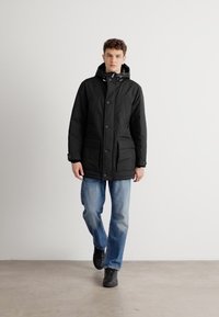 Črna izolirana jakna s kapuco, sprednjim gumbom in dvema velikima žepoma; nosi se čez svetlo modre jeans hlače in črne superge.