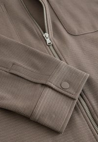 Manche de veste en tissu texturé taupe avec bouton de poignet et fermeture éclair argentée partiellement ouverte sur une poche avant assortie.