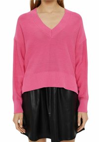 Różowy sweter z dekoltem w serek, zaszyty w luźnym stylu z opadającymi ramionami i prążkowanym dołem, w zestawieniu z czarną skórzaną spódnicą. Gładka tekstura i luźny krój.