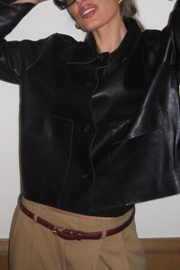 Čierna kožená skrátená bunda s dvoma prednými vreckami, s gombíkovým zapínaním a hladkou textúrou, nosená na béžových nohaviciach s hnedým opaskom.