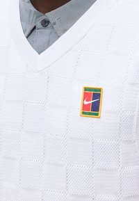 Pull blanc texturé à col en V porté sur une chemise grise à motifs avec des boutons, présentant un patch carré multicolore avec le logo Nike sur la poitrine.
