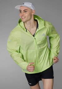 Leichte neongrüne Windjacke mit Kapuze, durchgehendem Reißverschluss und reflektierenden Akzenten. Getragen über einem schwarzen ärmellosen Top und Shorts.