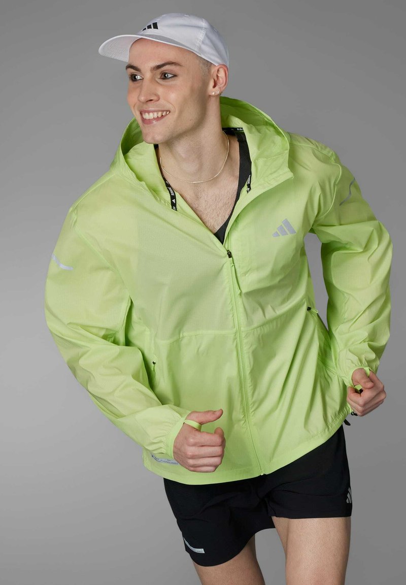 Leichte neongrüne Windjacke mit Kapuze, durchgehendem Reißverschluss und reflektierenden Akzenten. Getragen über einem schwarzen ärmellosen Top und Shorts.