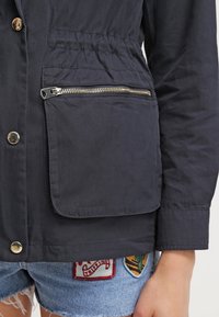 Navy jacket with a zippered pocket, silver hardware, and a drawstring waist. Smooth fabric, featuring gold buttons and detailed stitching.
