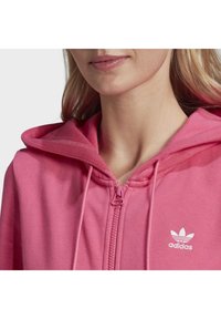 Růžová mikina na zip s velkou kapucí, vyrobená z měkké látky. Na levé straně hrudi je bílý logo Adidas a sladěný detail zipu.