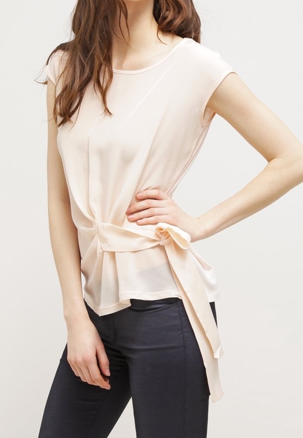 Femme portant un chemisier sans manches beige clair avec un détail noué sur le côté, associé à un pantalon sombre ajusté, posant avec une main sur la hanche.