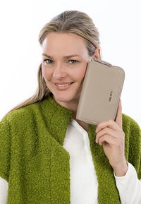 Lächelnde Frau mit blonden Haaren hält eine beige Reißverschluss-Geldbörse nah an ihr Gesicht, trägt einen strukturierten grünen Schal und ein weißes Hemd.