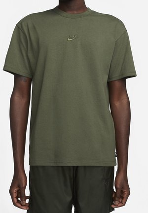 Mężczyzna w oliwkowozielonym t-shircie Nike z małym logo na piersi oraz czarnych szortach Nike, stojący z rękami swobodnie opuszczonymi wzdłuż ciała.