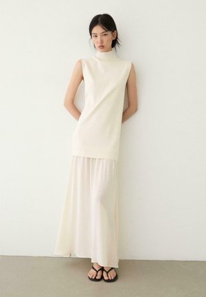 Frau in einem ärmellosen weißen Kleid mit hohem Kragen und einem fließenden Rock steht vor einer weißen Wand und trägt schwarze Sandalen.