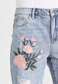Světle modré džínové kalhoty s potrháním, zdobené růžovou květinovou výšivkou a zelenými listy na levém stehně.