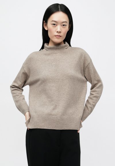 Ανοιχτό μπεζ πουλόβερ με ζιβάγκο και ελεύθερη εφαρμογή, με ριμπ μανσέτες και τελείωμα. Απαλή υφή με διακριτικό, ομοιόμορφο μοτίβο, φορεμένο πάνω από σκούρο παντελόνι.