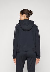 Svart hettejakke laget av lett materiale, med hvite aksentstriper langs skuldrene og en tettsittende elastisk midje.