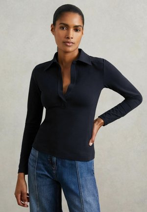Kvinde med mørkt hår trukket tilbage iført en tætsiddende sort langærmet skjorte med krave og blå jeans, stående mod en ensfarvet baggrund.