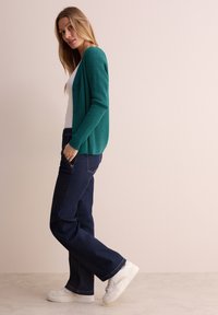Tropfengrüner, gerippter Cardigan mit Knöpfen, über einem weißen Oberteil getragen. Kombiniert mit dunkelblauen Jeans und weißen Sneakers, vor einem hellen Hintergrund präsentiert.