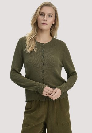 Frau mit langem blondem Haar, die eine olivgrüne, geknöpfte Strickjacke und passende Hosen trägt, steht vor einem einfarbigen hellen Hintergrund.