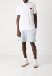 Polo blanc avec un écusson logo rouge, assorti à un short à rayures bleu clair. Des baskets blanches à semelle gomme complètent le look.