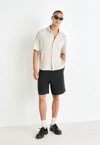 Lys beige ribbet kortærmet skjorte med lomme, parret med sorte shorts og sorte snørestøvler. Modellen har sorte solbriller og hvide sokker på.