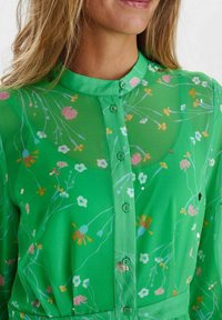 Grön, transparent blus med blommönster i rosa, gult och blått. Har knäppning framtill och krage, med subtil textur och figurnära midja.