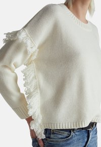 Biały sweter dzianinowy z fakturowanymi frędzlami na rękawach, prążkowanym dekoltem oraz luźnym fasonem, zestawiony z niebieskimi dżinsami.