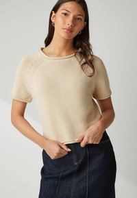 Ung kvinna med långt mörkt hår som bär en beige kortärmad stickad tröja och en mörk denimsböljande kjol, stående mot en neutral bakgrund.