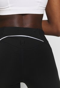 Sorte athletic leggings med høj talje, hvide accentstriber og en struktureret logo-detalje bagpå. Glat, strækbart stof.