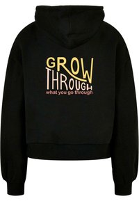Sudadera recortada negra hecha de tela suave. Cuenta con capucha y puños acanalados. Texto en el frente: "CRECE A TRAVÉS de lo que pasas" en letras coloridas.
