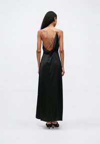 Femme aux cheveux bouclés portant une longue robe dos nu en satin noir avec de fines bretelles croisées, debout sur un fond blanc uni.