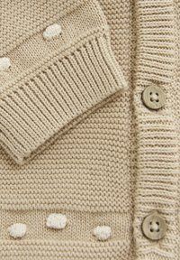 Beige gestrickte Strickjacke mit geripptem Bündchen, strukturiertem Muster mit weißen Bobbels und Holzknöpfen entlang der Vorderseite.