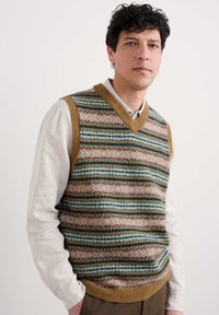 Uomo con capelli ricci e scuri che indossa un gilet a maglia a righe multicolore sopra una maglietta a maniche lunghe beige, su uno sfondo chiaro e uniforme.