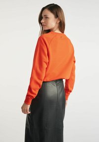 Pomarańczowa bluza z okrągłym dekoltem i rękawami raglanowymi, w połączeniu z czarną skórzaną spódnicą midi o gładkiej fakturze.
