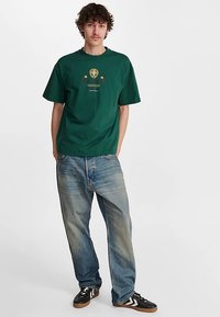 Groen katoenen t-shirt met een ronde hals en korte mouwen, voorzien van een centraal grafisch ontwerp. Gecombineerd met losse, vervaagde blauwe jeans en zwarte sneakers.